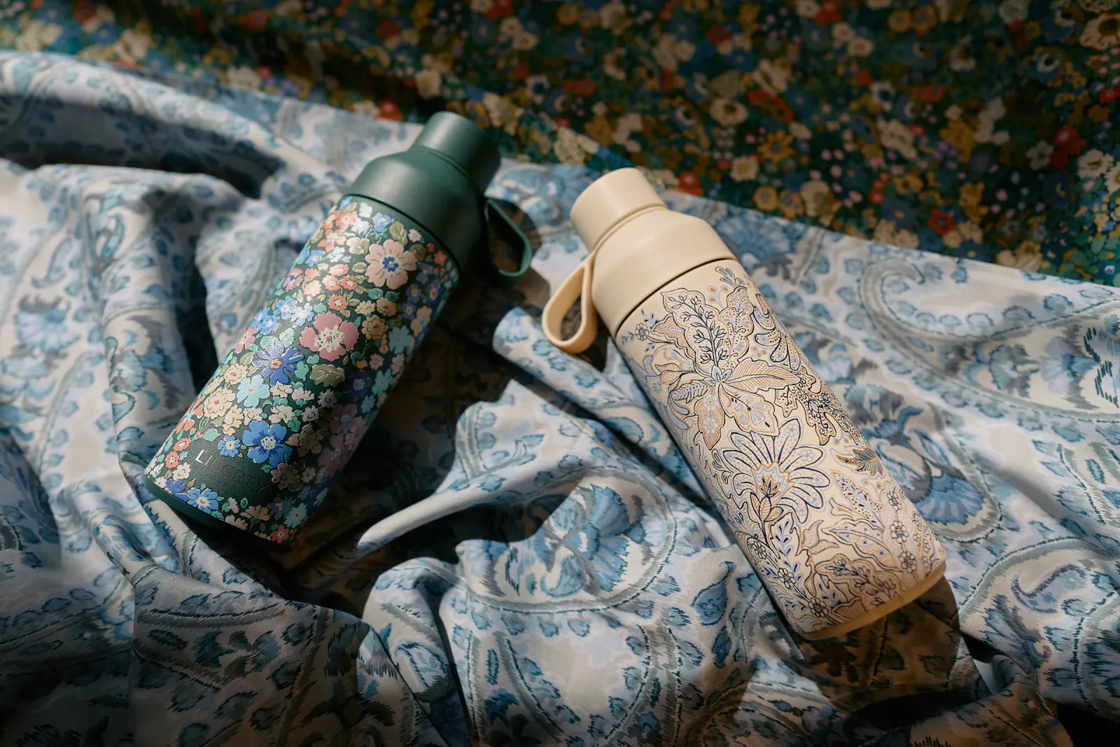 Two floral-patterned reusable water bottles on blue paisley fabric, one colorful and one beige