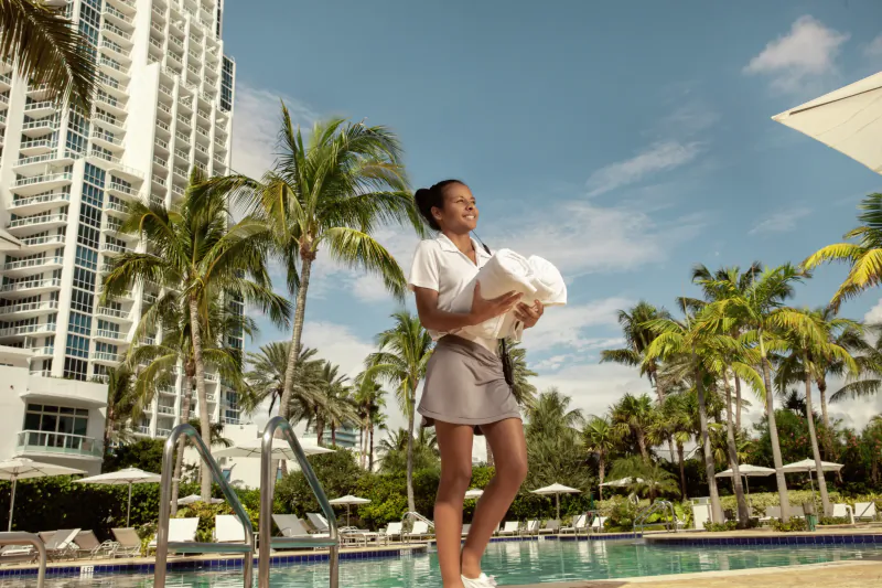 Smiling woman in white polo and grey skirt holding towels poolside at luxury Miami hotel with palm trees and high-rise