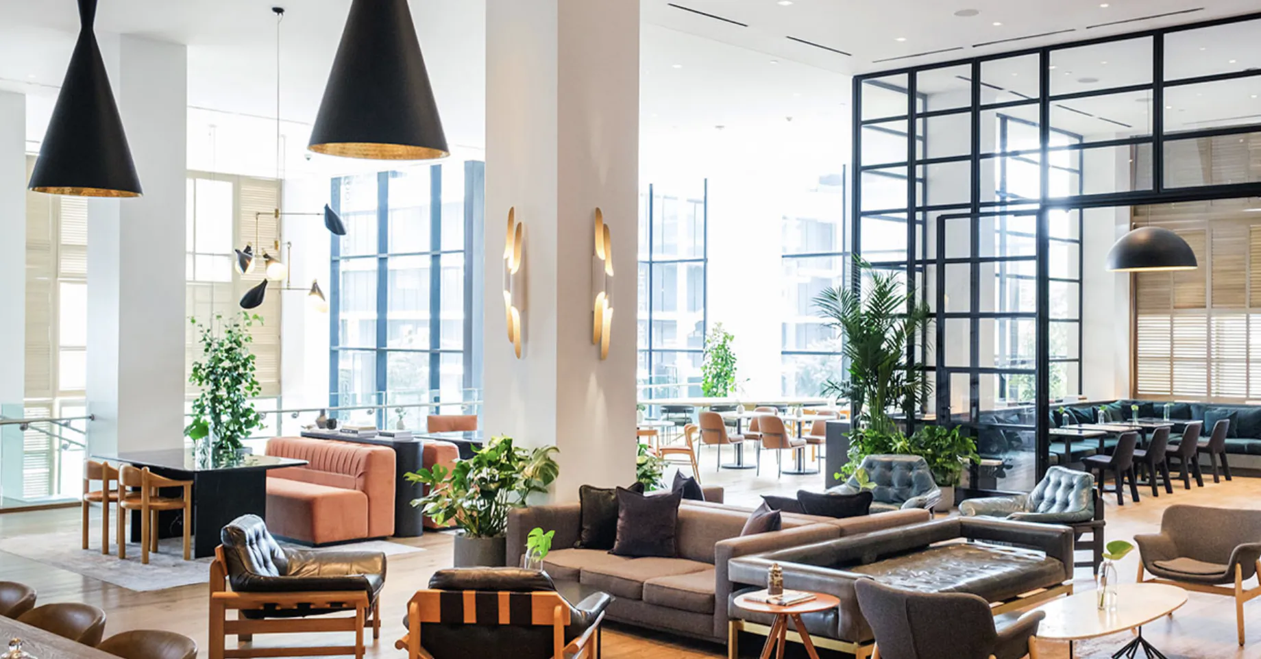 Modern hotel lobby with grey sofas, orange armchairs, black pendant lights, and large windows overlooking a dining area