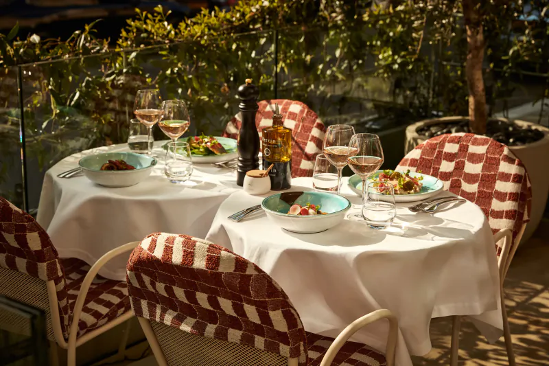Outdoor restaurant table set for four with white tablecloth, red checkered chairs, wine glasses, and plated salads