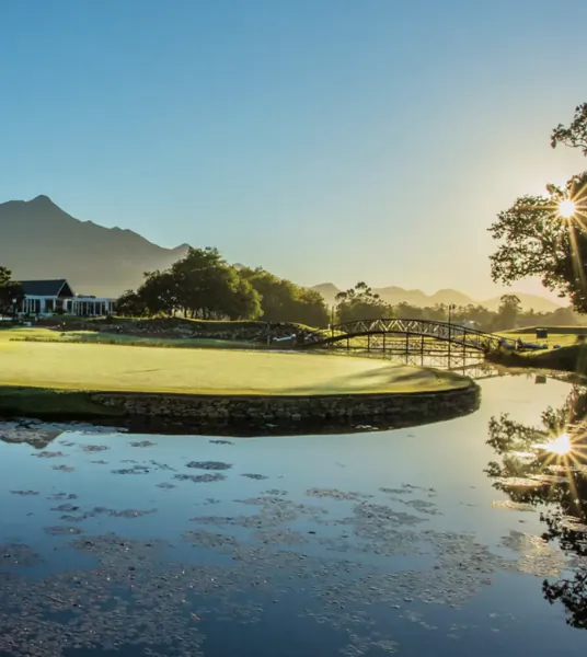 Golf course green with stone bridge over pond at sunrise, mountains and clubhouse in background