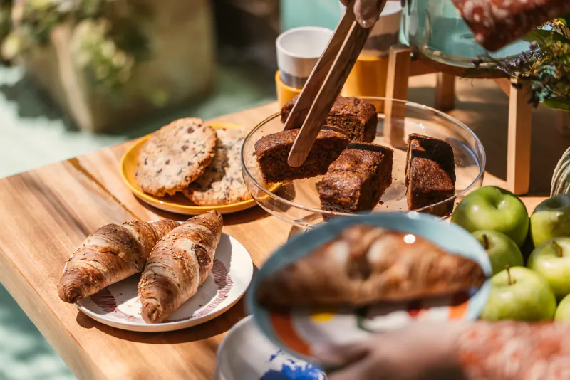 Hands using tongs to serve chocolate brownies from a glass dish on a wooden table with croissants, cookies, green apples, and a coffee cup