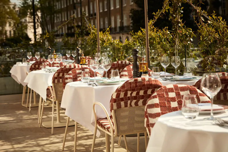 Outdoor restaurant patio with white tablecloths, red checkered chairs, and neatly set tables with wine glasses and napkins