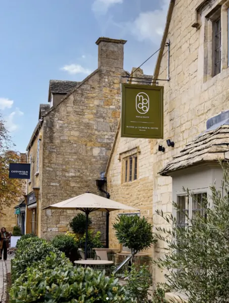 Stone Cotswold building with green 'The Cotswold House' sign, outdoor seating and umbrella
