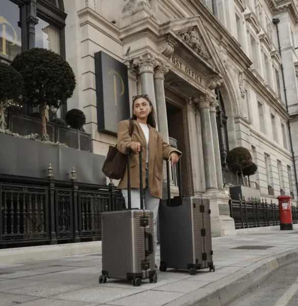 Woman in tan blazer stands with two grey suitcases outside a historic Post Office building