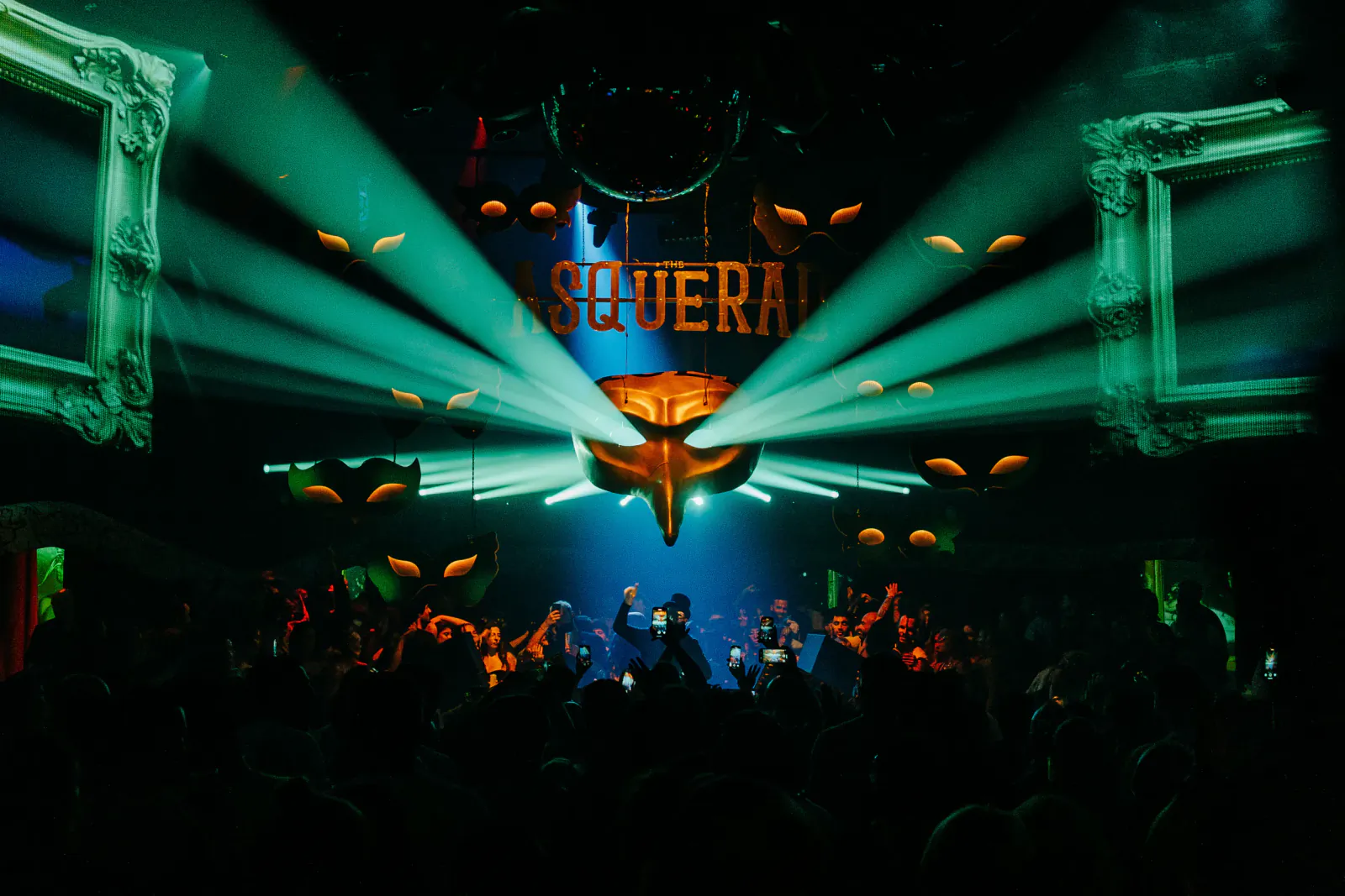 Crowd dancing under green spotlights at a nightclub with 'ASQUERAI' sign and ornate frames
