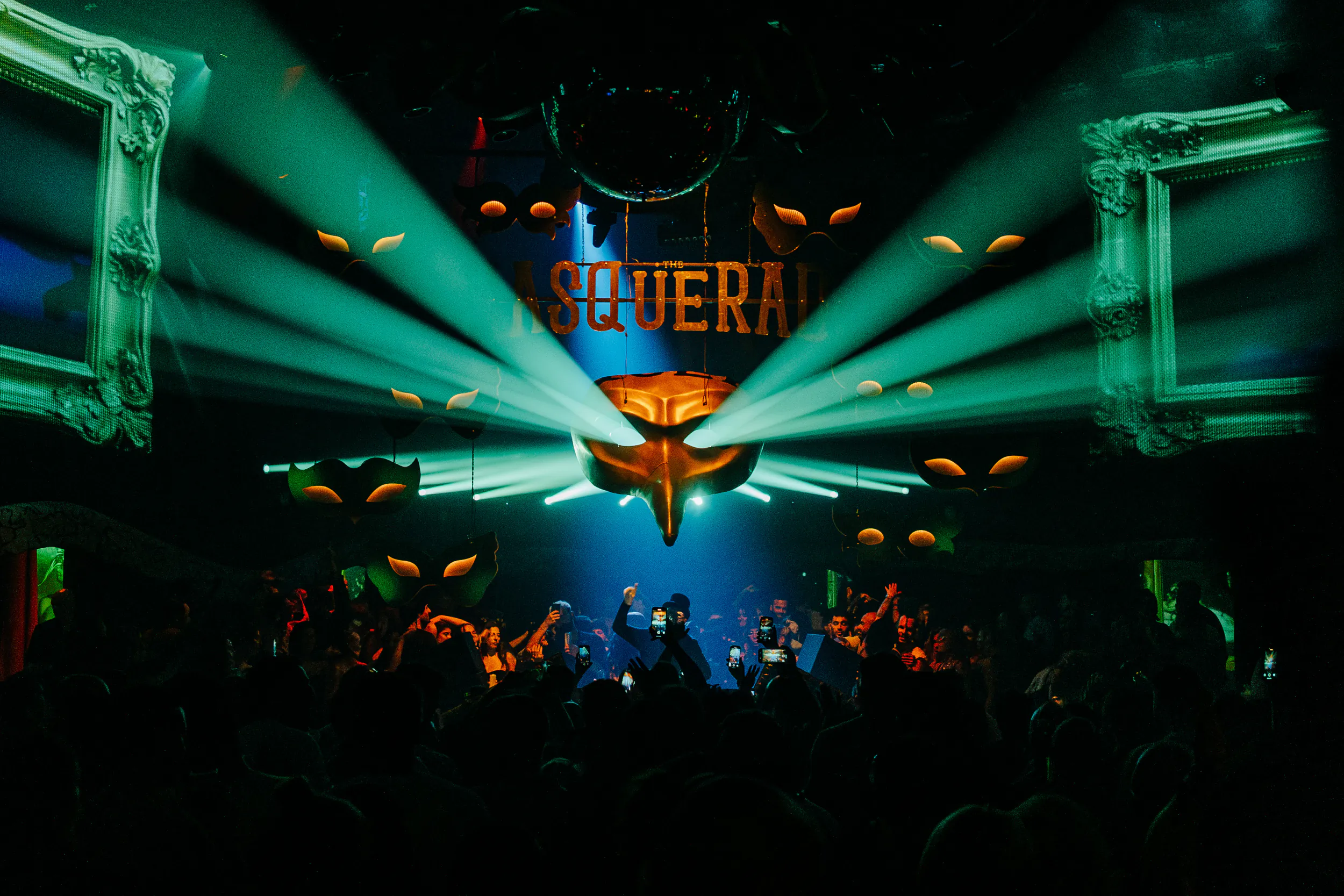 Crowd dancing under green spotlights at a nightclub with 'ASQUERAI' sign and ornate frames