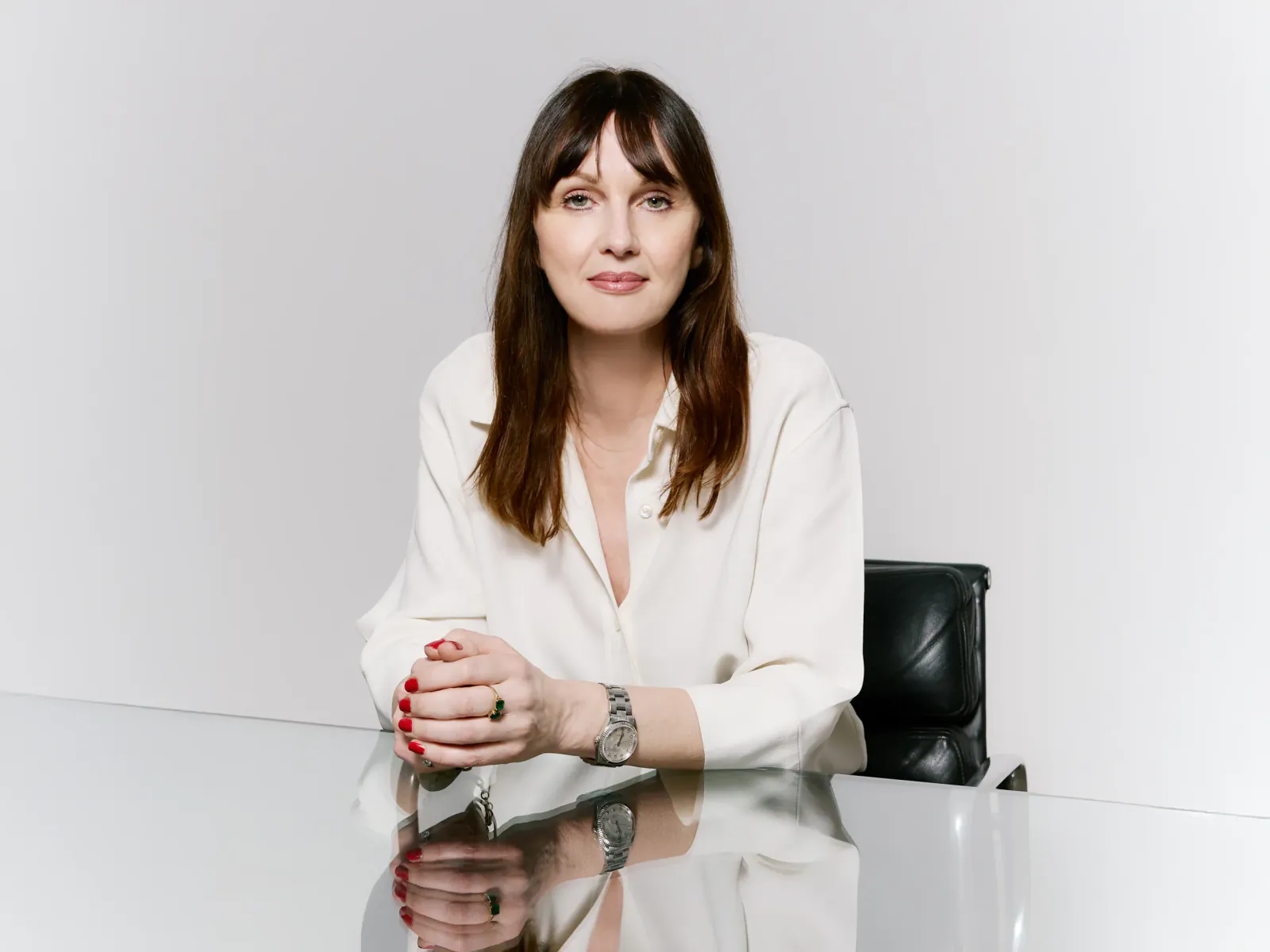 Woman with long brown hair in white shirt seated at glossy table, hands clasped, wearing watch and red nail polish