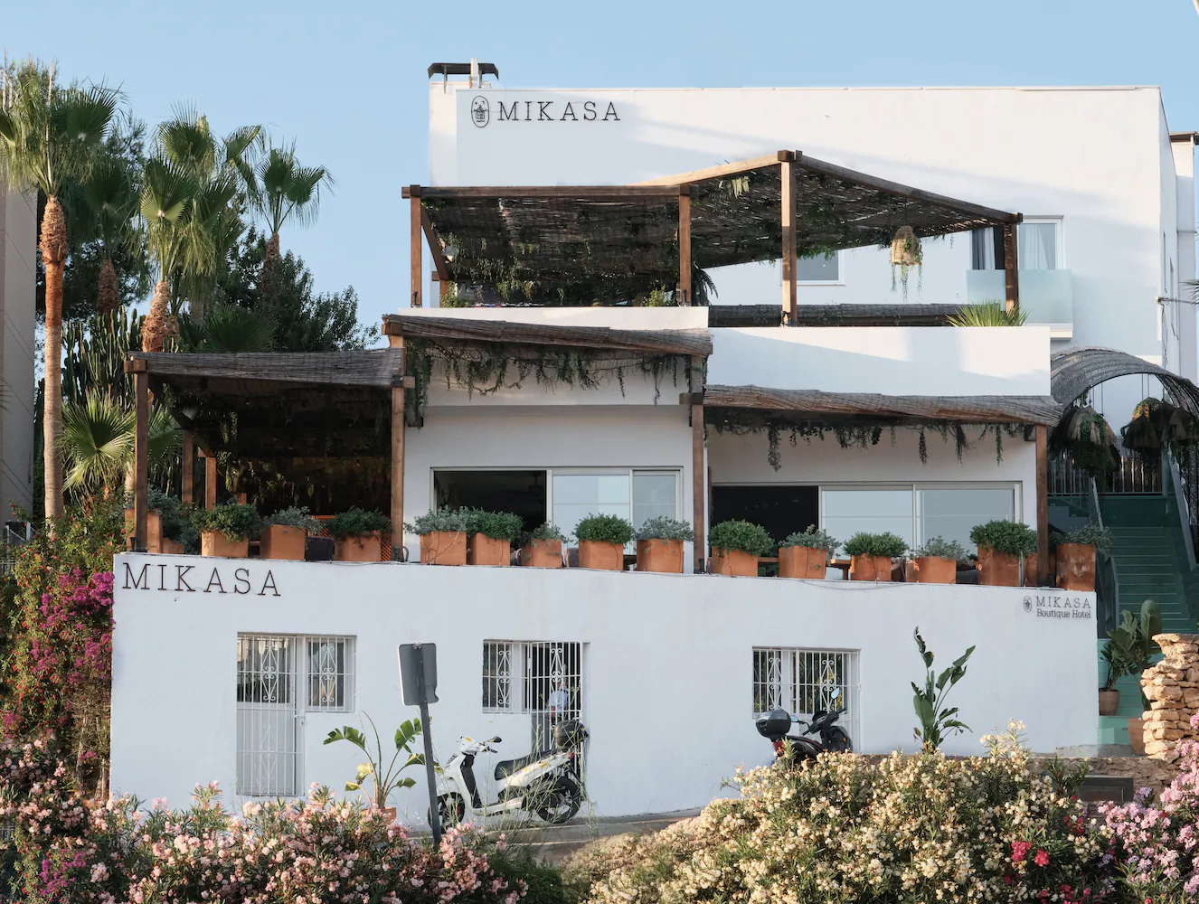 White Mikasa building with terracotta planters, scooters parked outside, and palm trees under clear sky