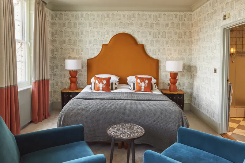 Hotel bedroom with orange headboard, lobster pillows, blue velvet chairs, and patterned wallpaper