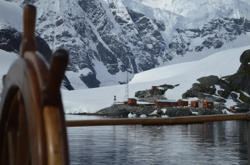 Ship's wooden wheel overlooking red research station buildings on snowy Antarctic peninsula with icy mountains