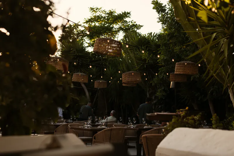 Outdoor restaurant patio at dusk with woven pendant lights, string lights, and diners seated among lush greenery