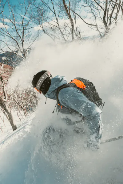 Snowboarder in blue jacket and black helmet kicking up powder on a snowy slope with bare trees