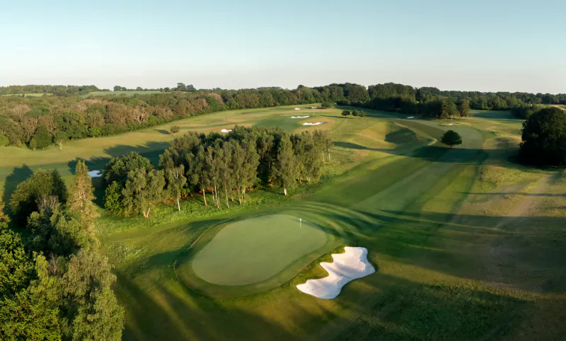 Aerial view of a golf course green with sand bunker and flag, surrounded by trees and fairways