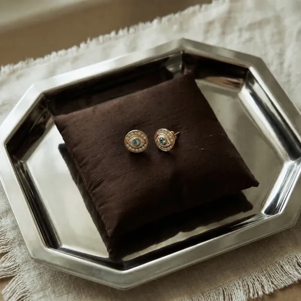 Two gold stud earrings with turquoise stones and diamond halos rest on a dark brown silk pillow inside a polished silver octagonal tray