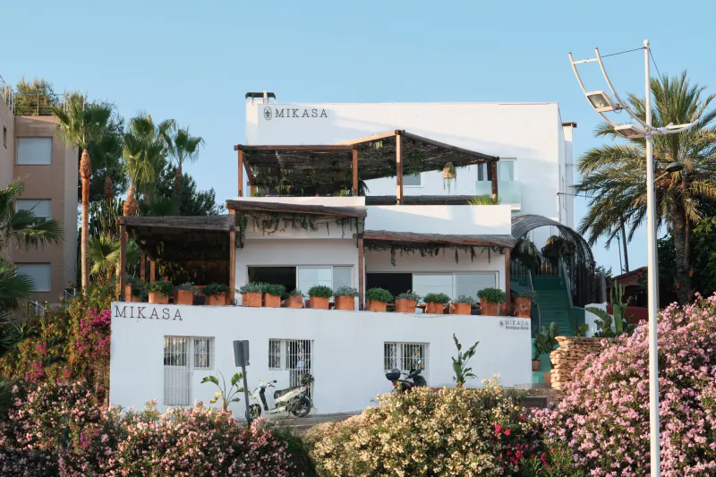 White Mikasa building with terracotta pots, scooter parked outside, palm trees and pink flowers in Ibiza