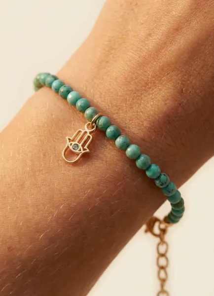 Close-up of a wrist wearing a turquoise beaded bracelet with a gold Hamsa hand charm featuring a small blue gem