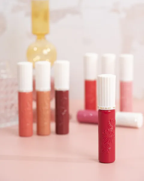 Paul & Joe lip gloss tubes in various shades on pink surface with yellow vase
