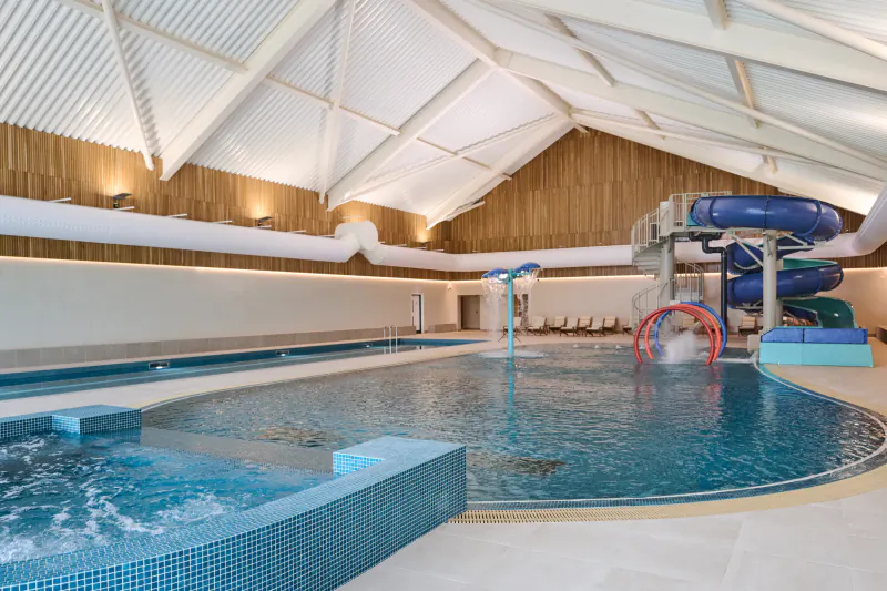 Indoor leisure pool with blue water slides, fountain, and hot tub in a modern spa with white vaulted ceiling