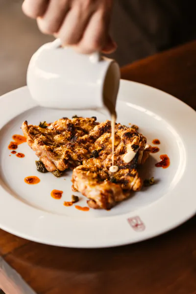 Hand pouring creamy sauce from a white jug over grilled pork chop on white plate with red sauce dots