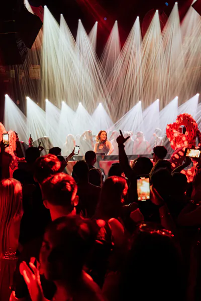 Female performer on stage under bright white spotlights, audience holding up phones and hands, red stage lights and dramatic lighting at Chinois Ibiza