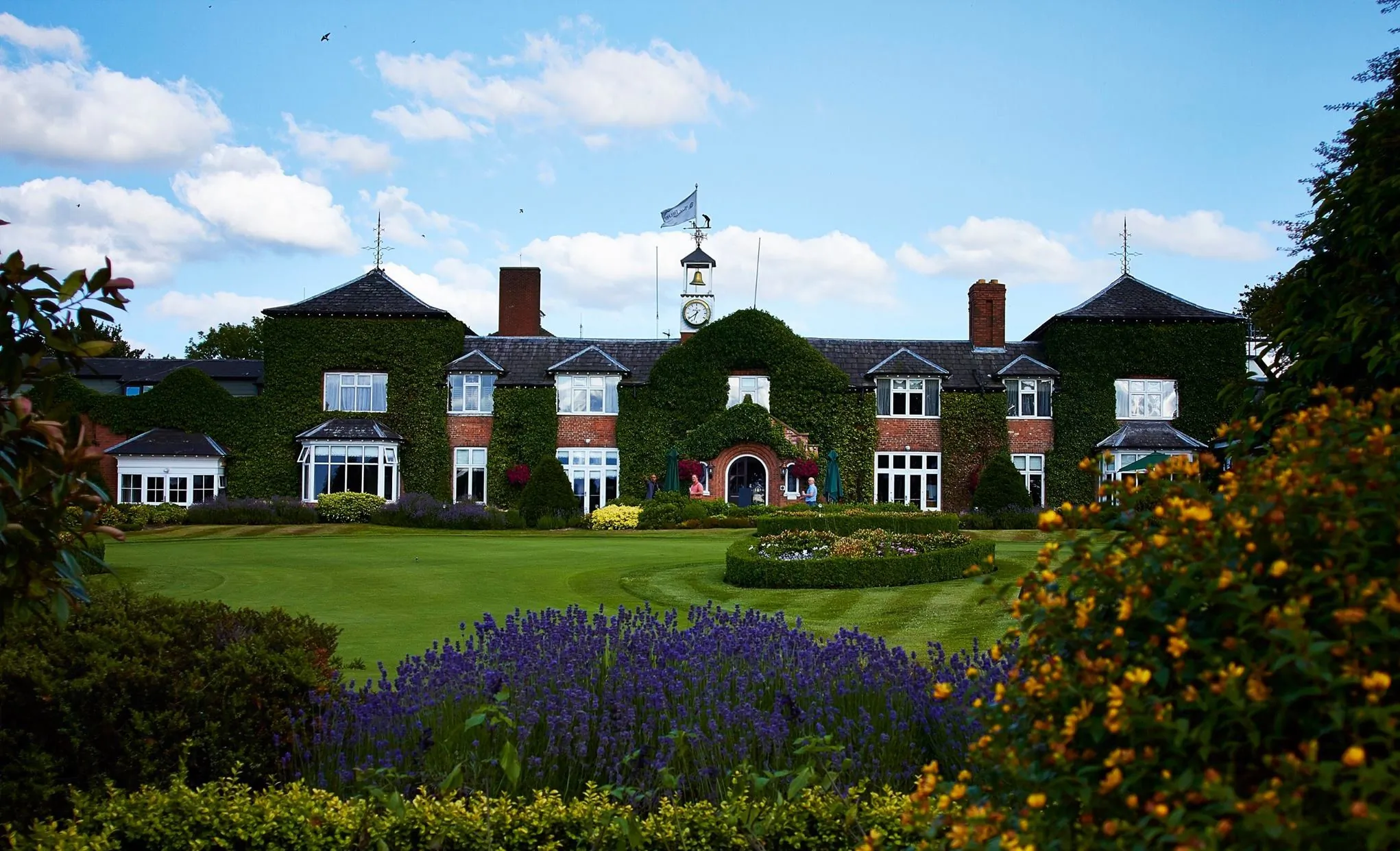 The Belfry hotel with ivy-covered brick facade, clock tower, and flag, surrounded by manicured lawns and colorful flower gardens under a blue sky