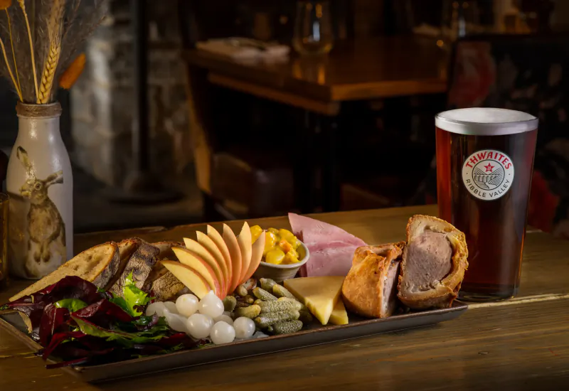 Ploughman's platter with sliced meats, cheeses, pickles, salad and pint of Hawkes Triple Tipple beer on wooden pub table