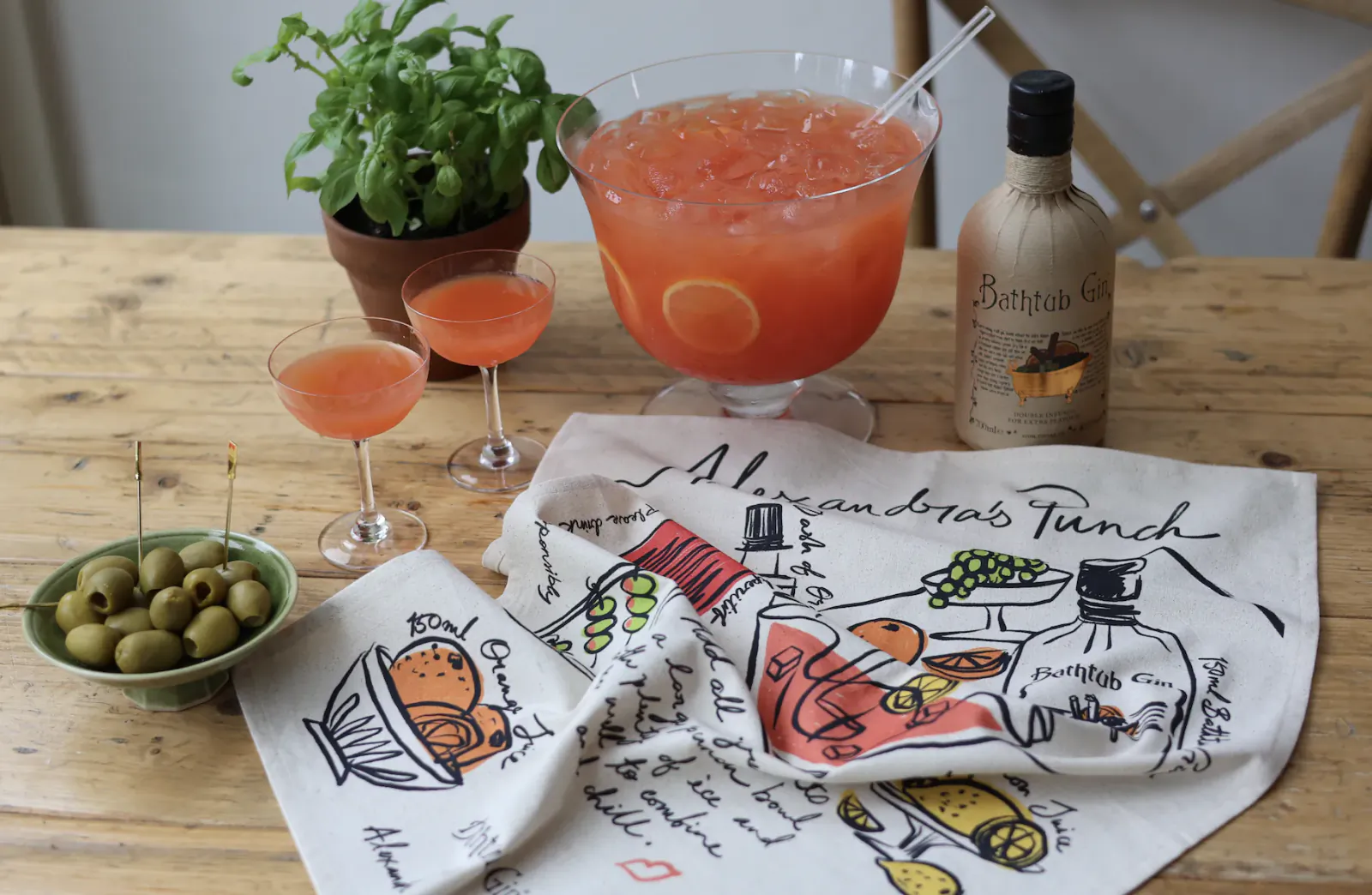 Wooden table with large glass punch bowl of red-orange cocktail, two filled coupe glasses, Bathtub Gin bottle, bowl of green olives, potted basil plant, and Alexandra's Punch illustrated tea towel