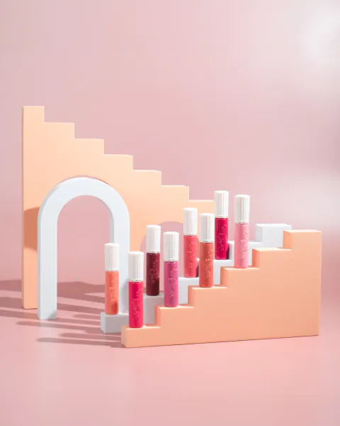 Assorted pink and coral lip gloss tubes arranged on peach geometric stairs with an arch backdrop