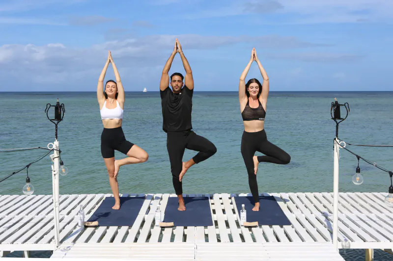 Three people holding tree pose on a wooden deck over the ocean, hands in prayer position above their heads