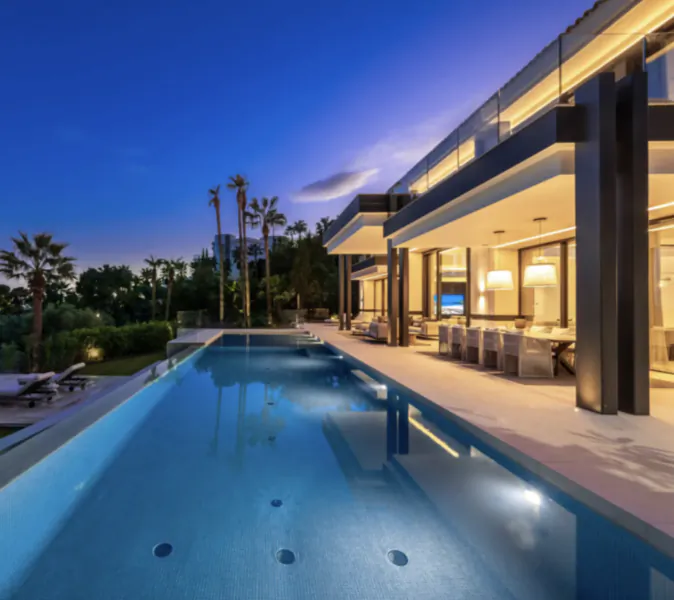 Modern luxury villa with long illuminated pool at dusk, open-plan living area with dining table and palm trees
