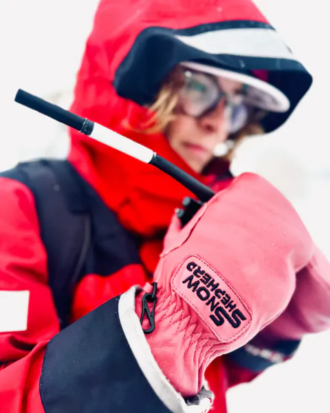 Person in red snow jacket and pink gloves holding a ski pole, close-up of SnowShepherd logo on glove