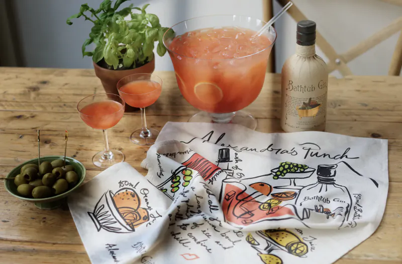 Wooden table with gin punch bowl, two glasses, Bath-tub Gin bottle, olives, basil plant, and illustrated napkins