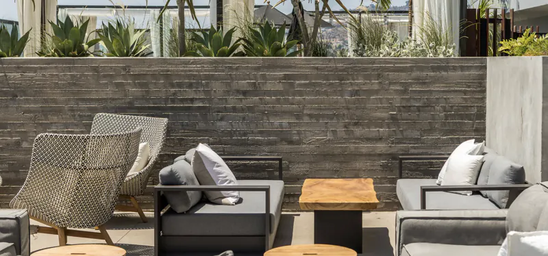 Modern outdoor patio with grey sofas, woven chairs, and a wooden coffee table against a grey brick wall with plants