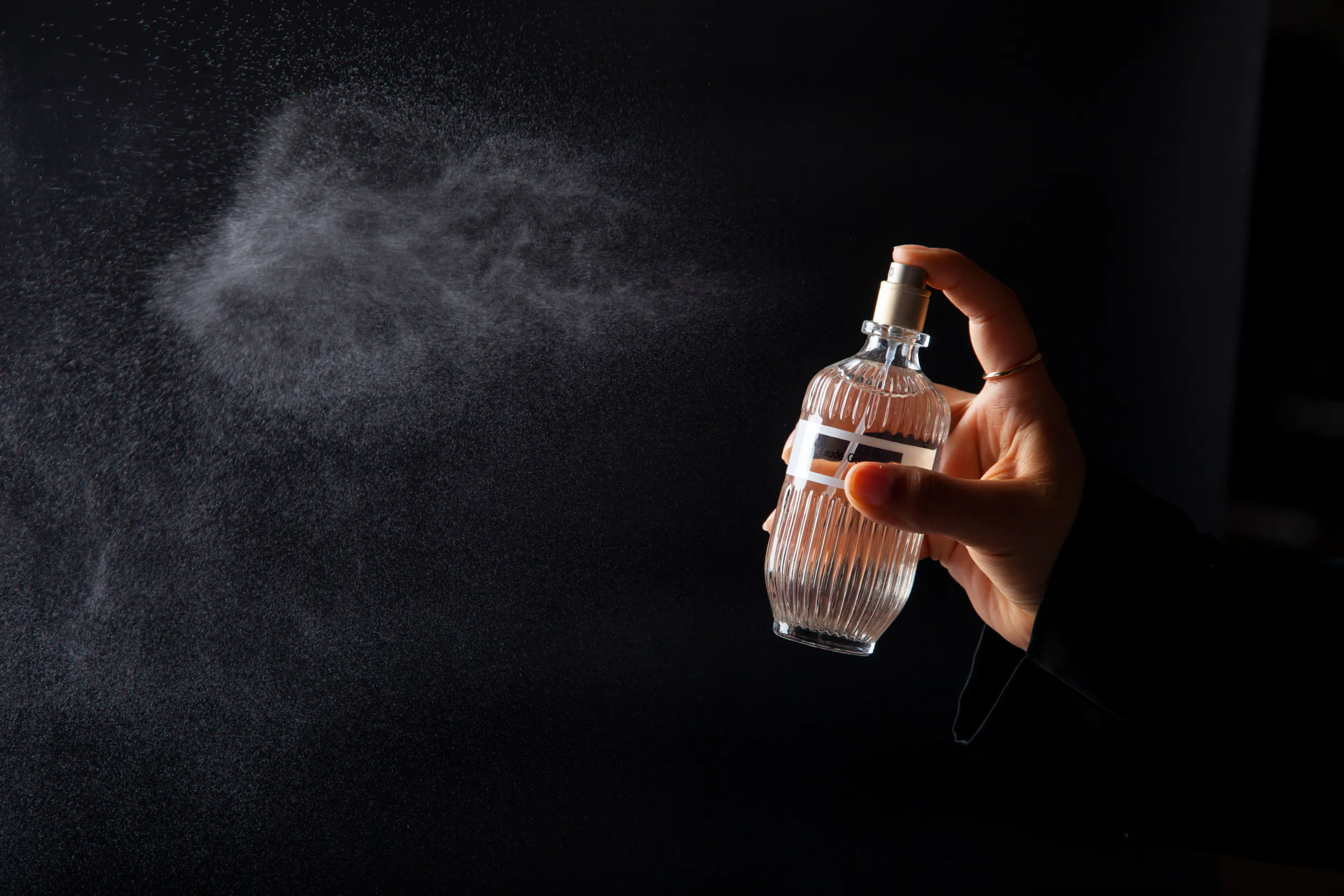 Hand spraying a clear glass perfume bottle, mist visible against black background