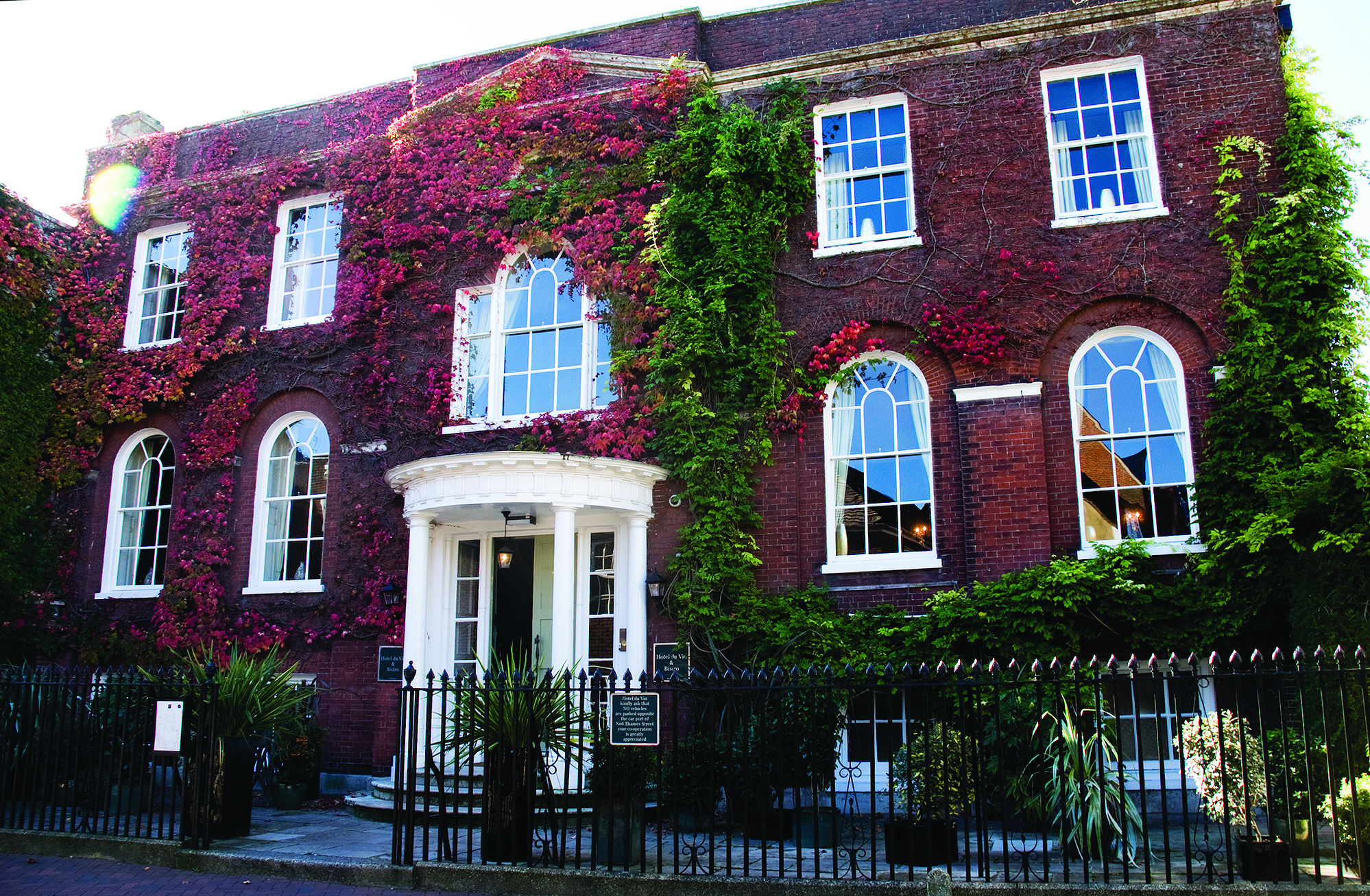 Red brick historic building covered in vibrant red and green ivy, with white arched windows and a central white portico entrance behind a black iron fence