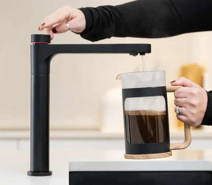 Person pressing button on black coffee maker as hot coffee pours into glass French press carafe