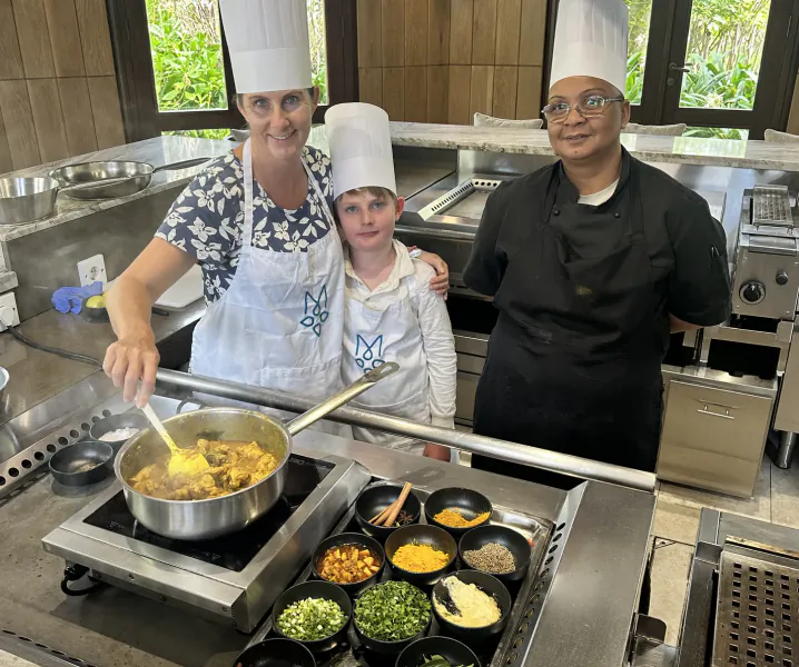 Woman and boy in chef hats stir curry in a pan while a male chef stands beside them in a professional kitchen