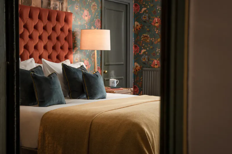 Hotel bedroom with burnt-orange tufted headboard, navy pillows, mustard throw, floral wallpaper and a steaming cup on the bed