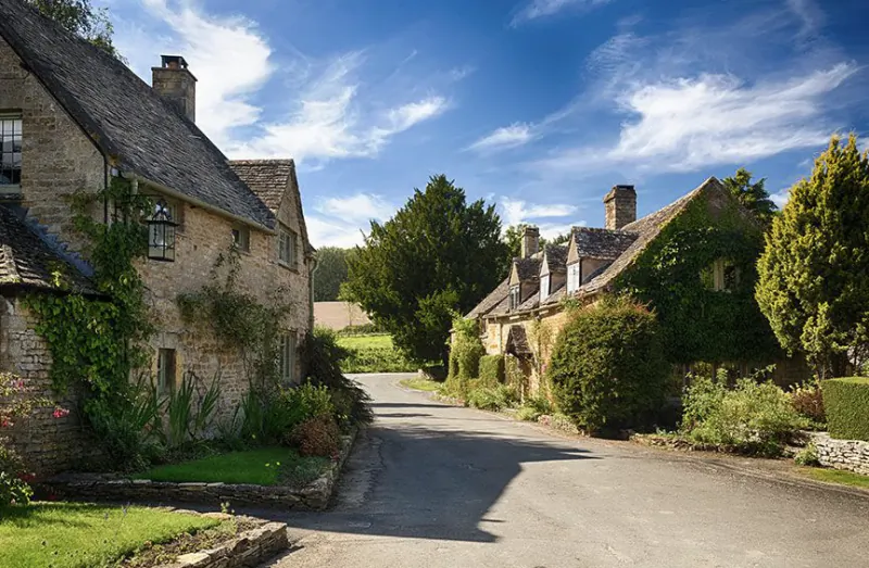Quiet country lane lined with ivy-covered Cotswold stone cottages under a bright blue sky