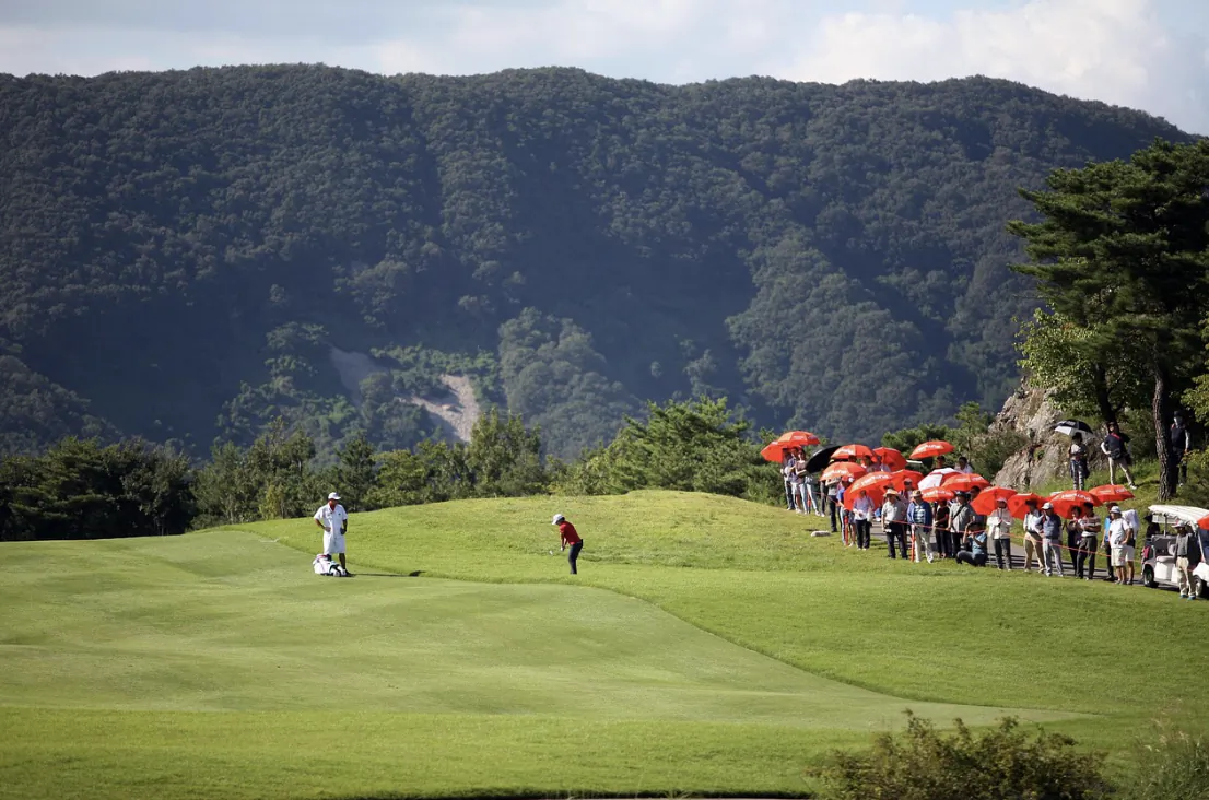 Golfer in red shirt tees off on lush green course with spectators holding red umbrellas and forested mountains behind