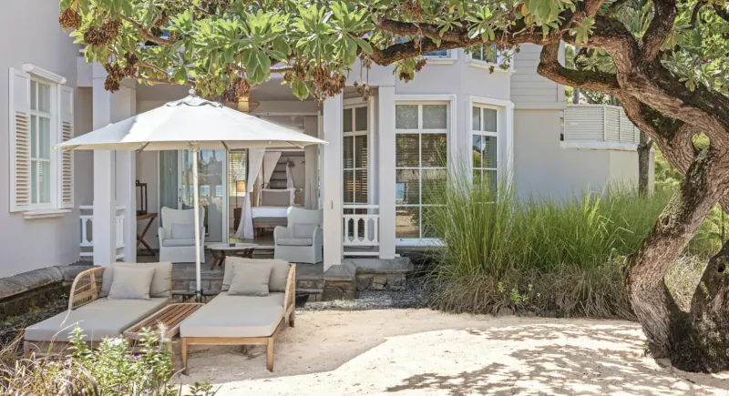 Luxury villa patio with two lounge chairs, white umbrella, and outdoor seating area under a large tree