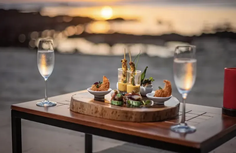 Sunset beachside table with two champagne glasses, assorted gourmet appetizers on wooden board, and red candle