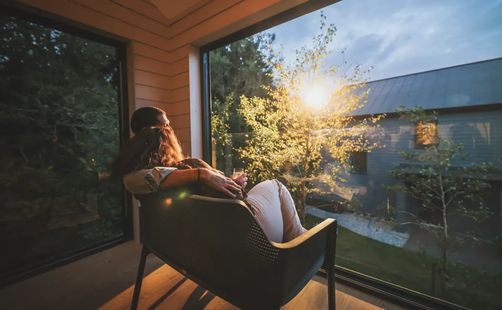 Woman with long hair relaxes in a chair by a large window, gazing at a sunlit tree outside at dusk