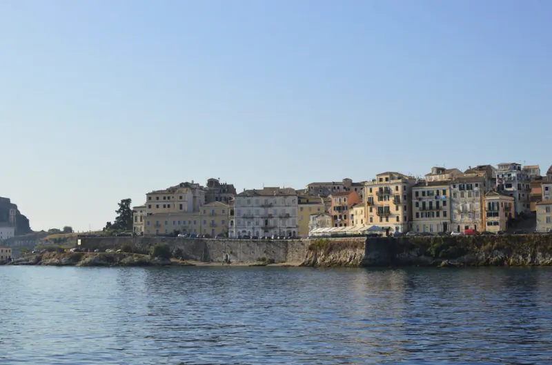 Colorful waterfront buildings line a rocky shore under a clear blue sky, viewed across calm water