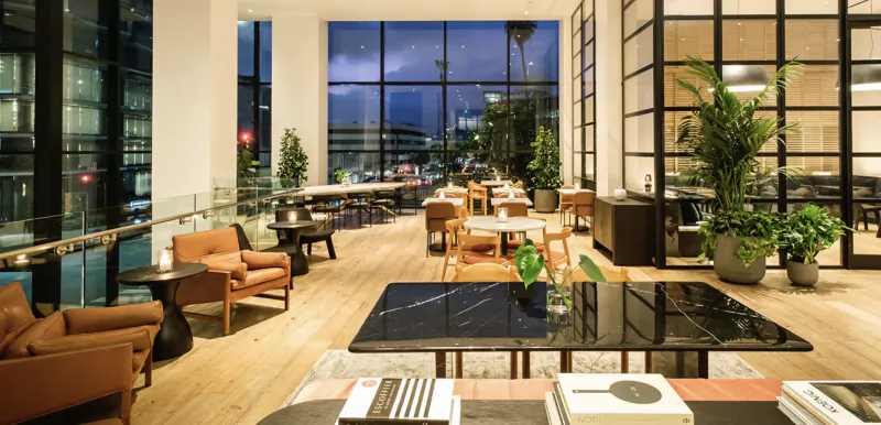 Modern hotel lobby lounge with leather armchairs, large black coffee table, and floor-to-ceiling windows overlooking city lights at dusk