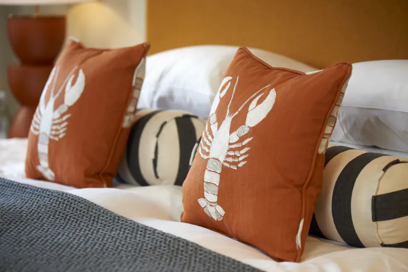 Two rust-orange pillows with white embroidered lobsters on a neatly made hotel bed with striped cushions