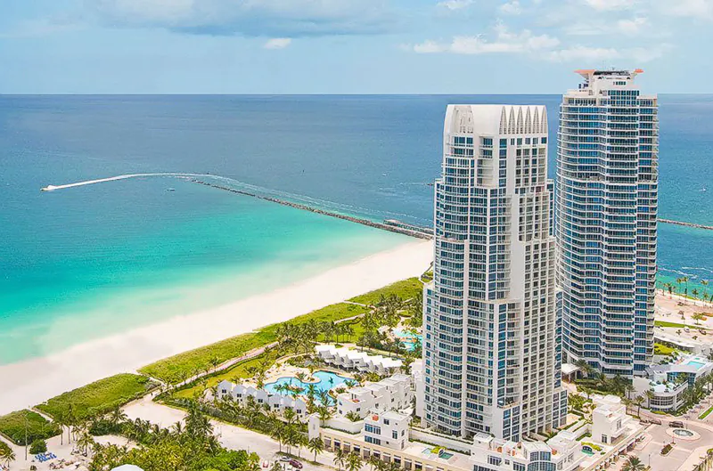 Aerial view of two high-rise luxury towers beside turquoise ocean and white sand beach in Miami Beach