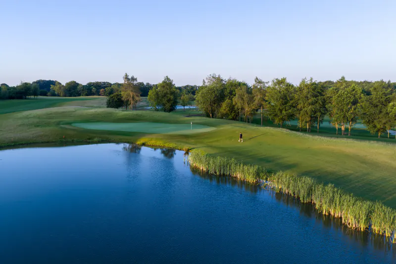 Golfer walking on a lush green fairway beside a calm blue lake with trees and a sand bunker in the background