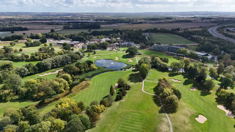 Aerial view of The Belfry golf resort showing lush fairways, a lake, hotel buildings, and surrounding countryside